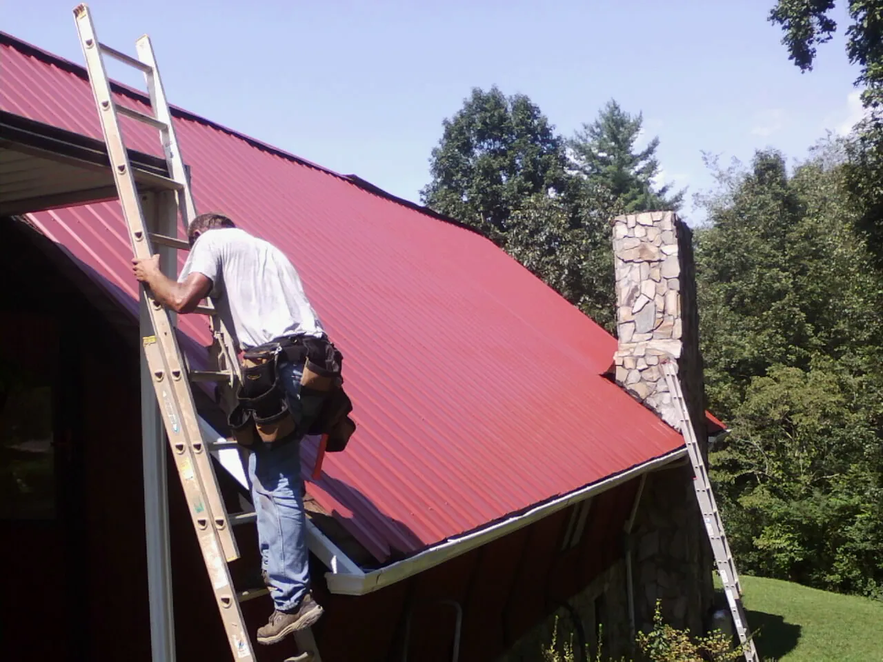 Roofer on a ladder beside a red metal roof and stone chimney — Soffit & Fascia Repair by Roofing Brothers in Bechtelsville